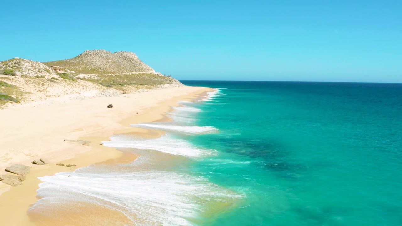 East Cape BCS Mexico coast with curling white surf near the lower left, a long empty-looking sand stretch, turquoise water filling the right side, and low rocky hills behind the beach.