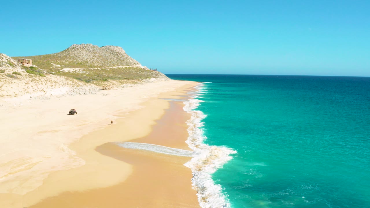 Aerial frame over East Cape BCS Mexico showing a sandy beach, turquoise ocean, white surf, a small 4x4 on the sand, and one person near the shoreline.
