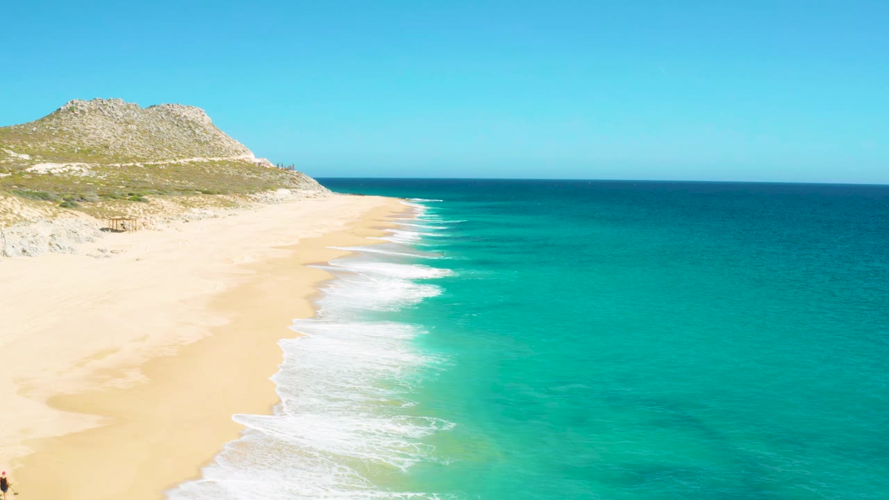 East Cape Baja California Sur shoreline with white wave bands moving up the sand, turquoise water on the right, rocky dune terrain on the left, and no visible people.