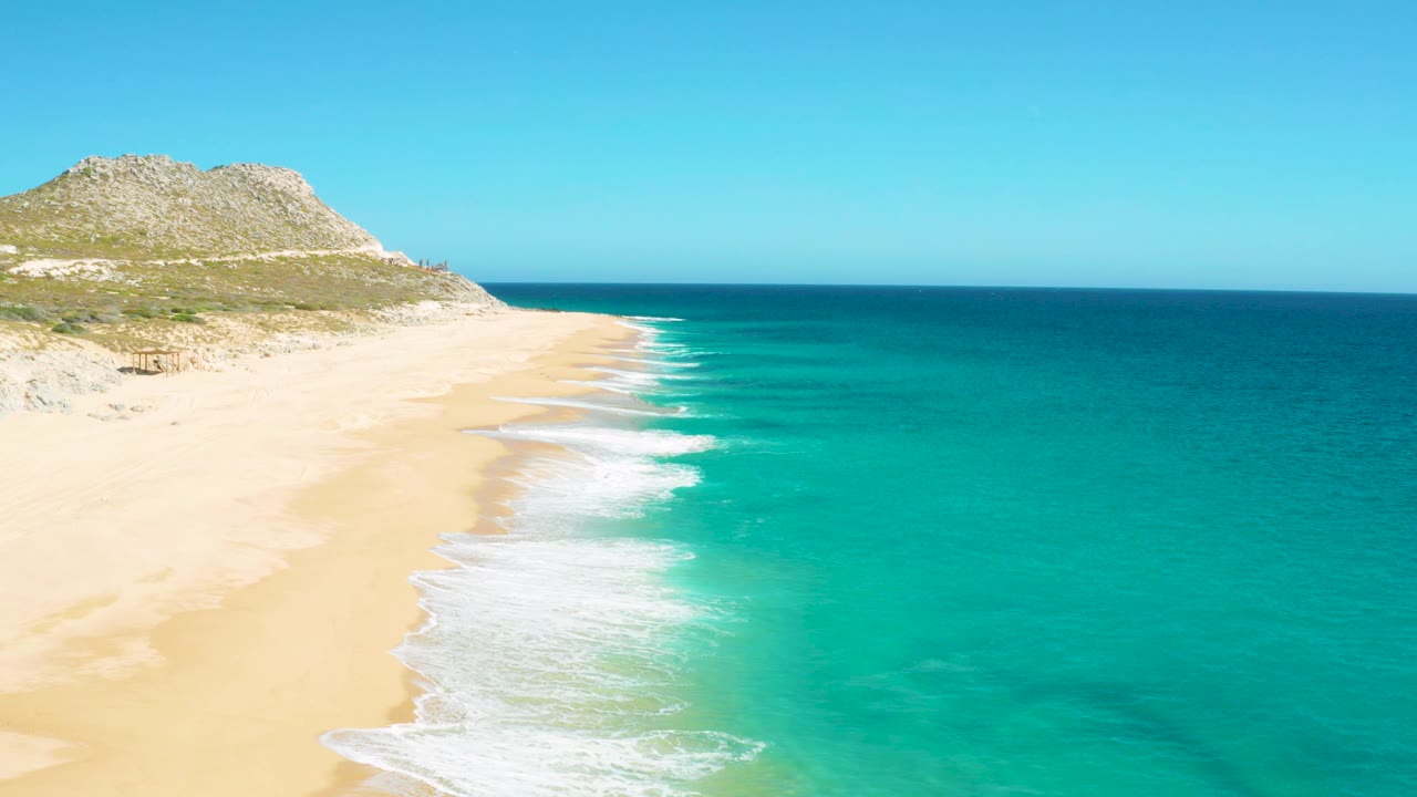 East Cape Baja California Sur beach with the surf line curling along the shore, deeper blue water near the horizon, rocky dunes to the left, and no visible people.