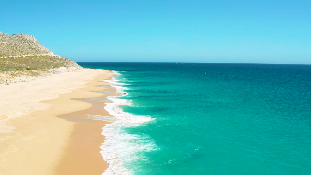 East Cape aerial view with white wave foam advancing along the sand and deeper blue water beyond the turquoise shallows, with no visible people.