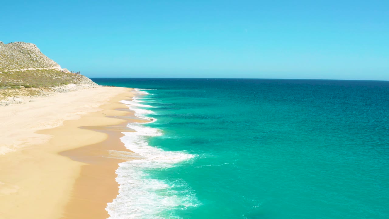 Aerial coastal frame in East Cape, Baja California Sur with a wider sweep of turquoise ocean on the right and surf curling onto empty sand at left, with no visible people.
