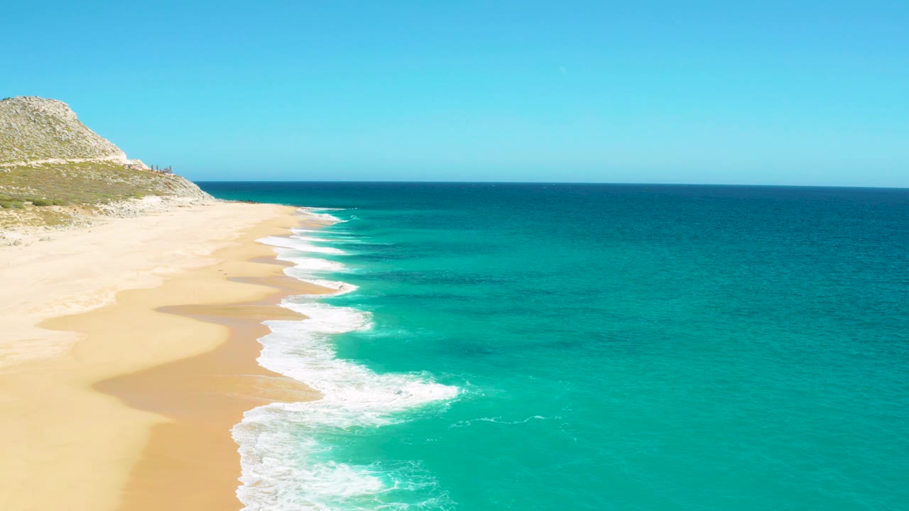 High coastal view of East Cape surf forming a bright foam edge beside pale sand, with the rocky slope at upper left and no visible people.
