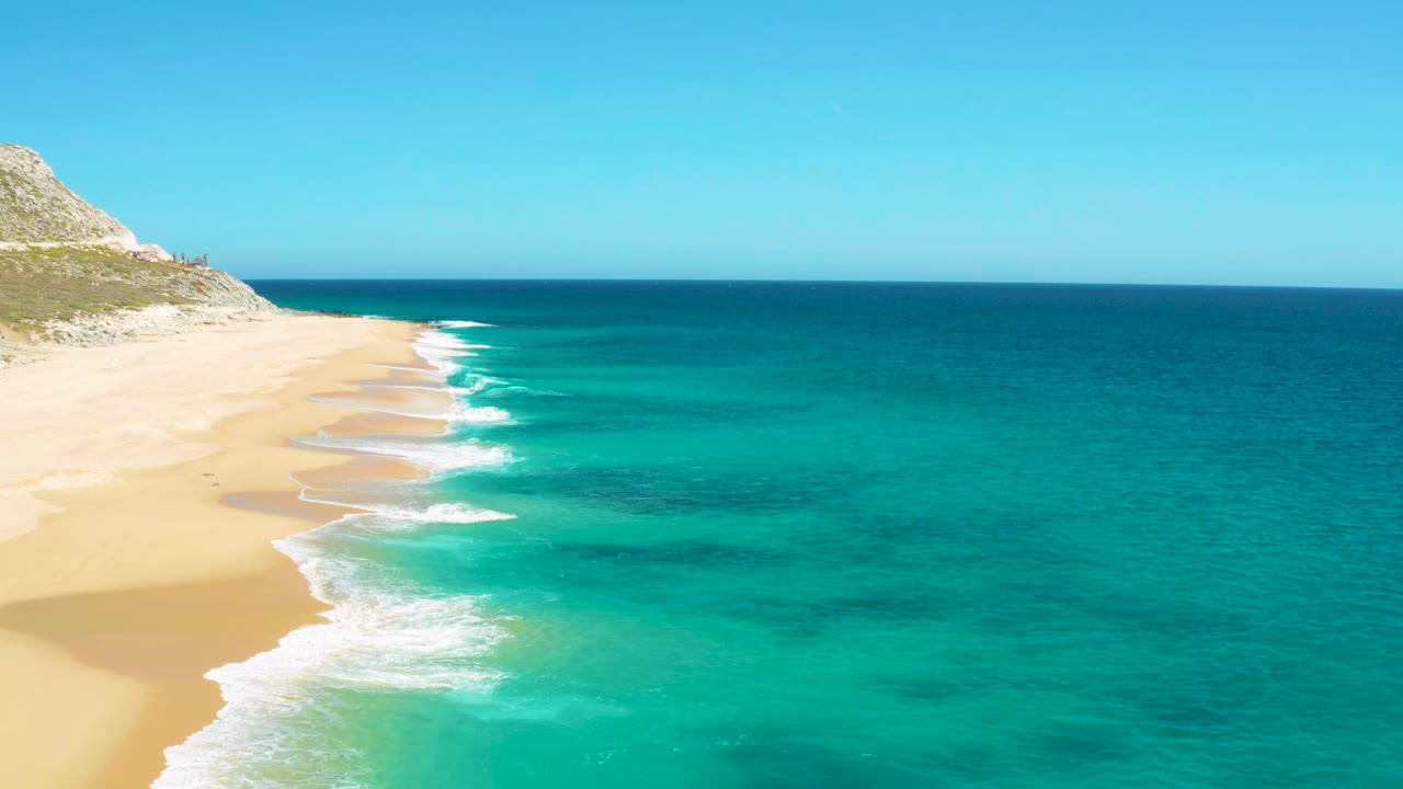 East Cape, Baja California Sur shoreline with repeating white wave breaks on tan sand, deeper blue water offshore, rocky slope at upper left, and no visible people.