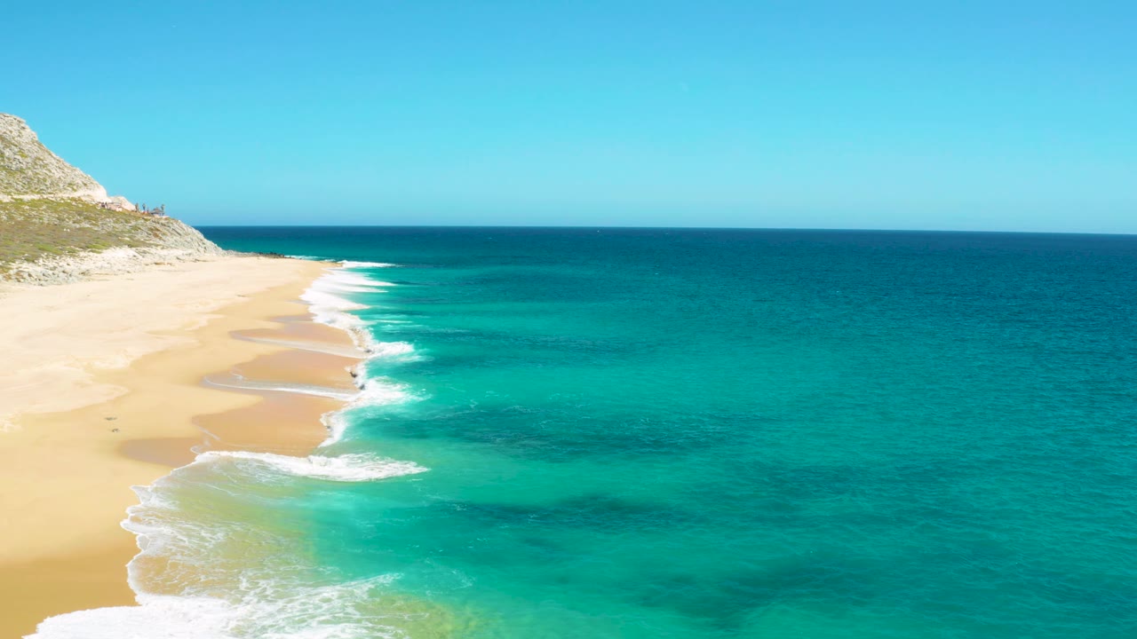 Mid-clip frame of East Cape, Baja California Sur with empty sand at left, a bright white surf edge, darker underwater patches in the turquoise sea, and no visible people.