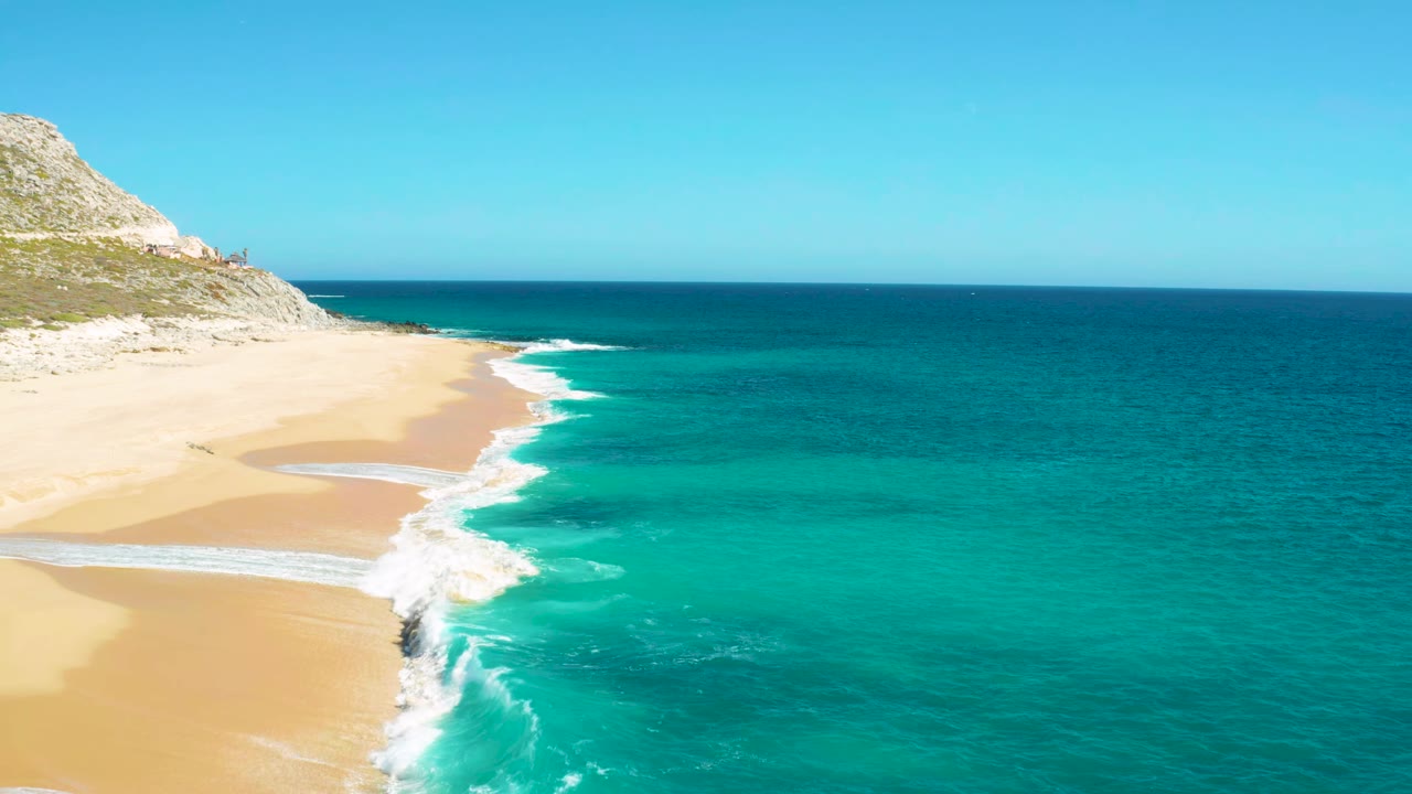 Turquoise surf breaks diagonally along a sandy beach under a clear sky.