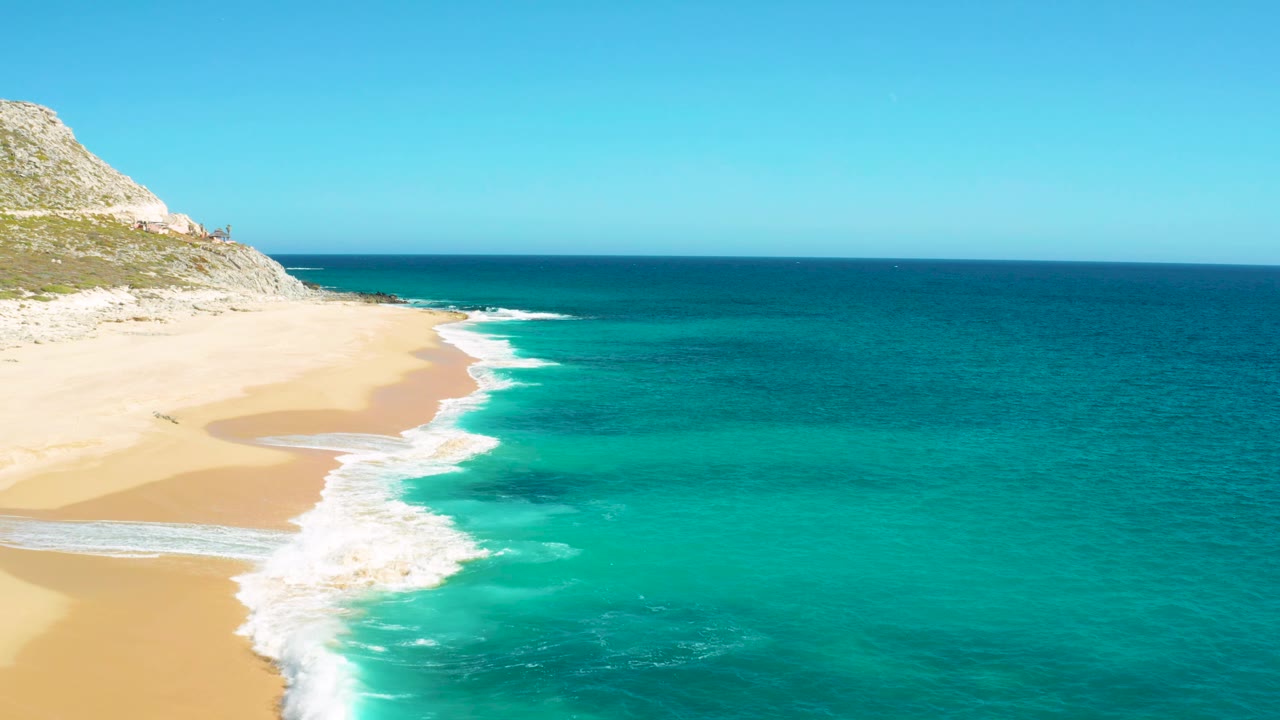 East Cape aerial frame with a wider patch of white surf foam near the lower left, open turquoise water to the right, and no visible people on the beach.