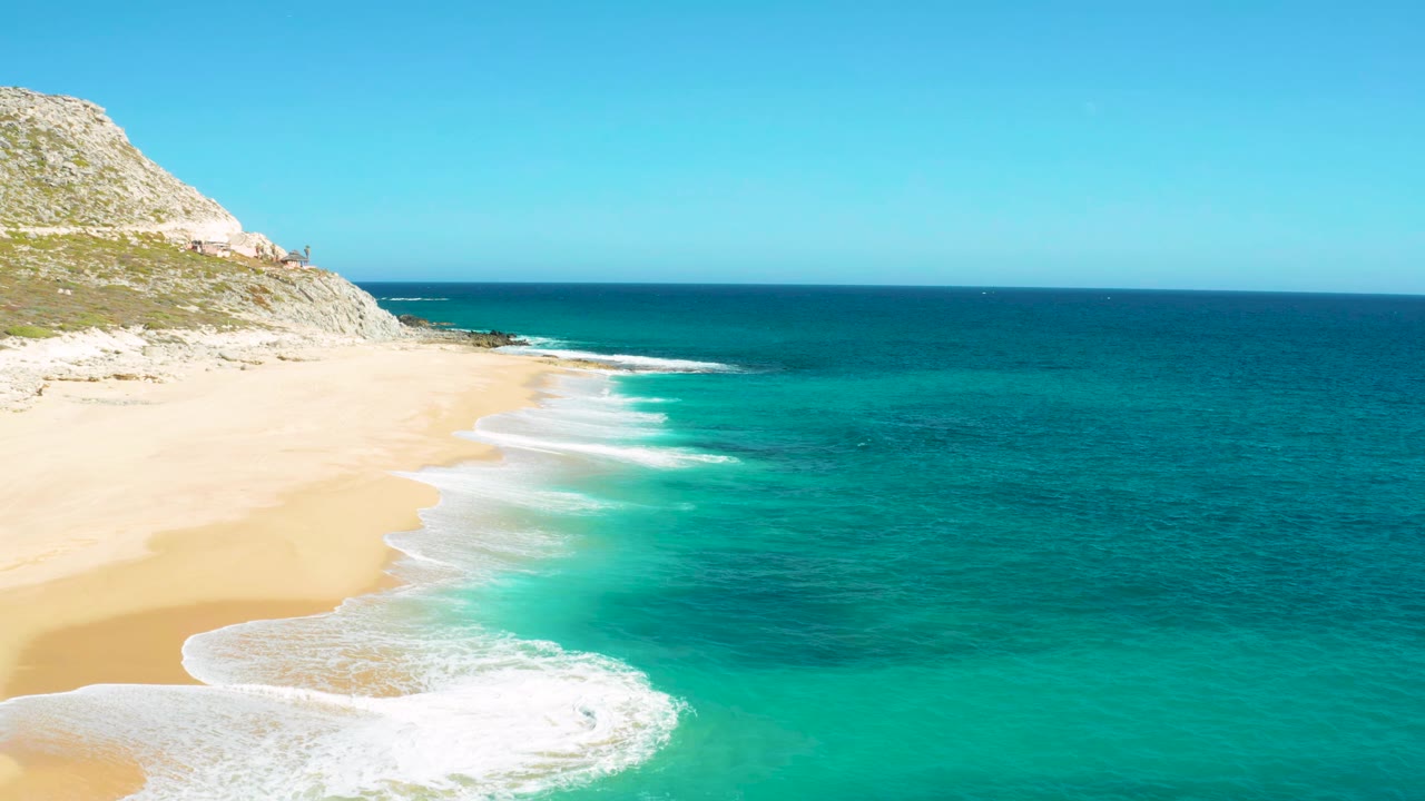 Aerial view in East Cape, Baja California Sur with the sandy beach widening at left, a white foam patch near the lower shore, turquoise water to the right, and no visible people.