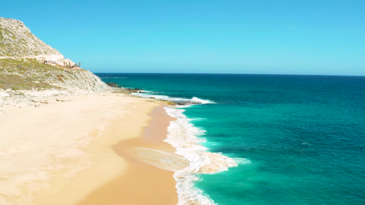 Aerial frame from East Cape, Baja California Sur showing pale sand, turquoise shore water, deeper blue ocean, a rocky cliff at left, and no visible people.