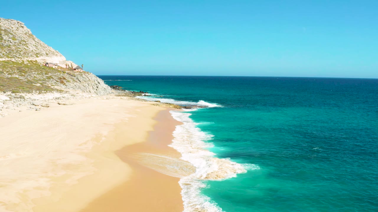 East Cape, Baja California Sur beach frame with a curved foam line advancing over turquoise shallows, pale sand at left, open ocean at right, and no visible people.