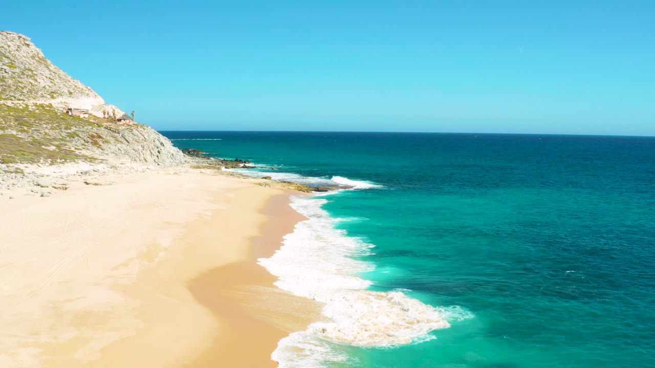 Aerial view in East Cape, Baja California Sur with the surf line shifted farther along the beach, a broad area of sand visible, rocky terrain at upper left, and no visible people.