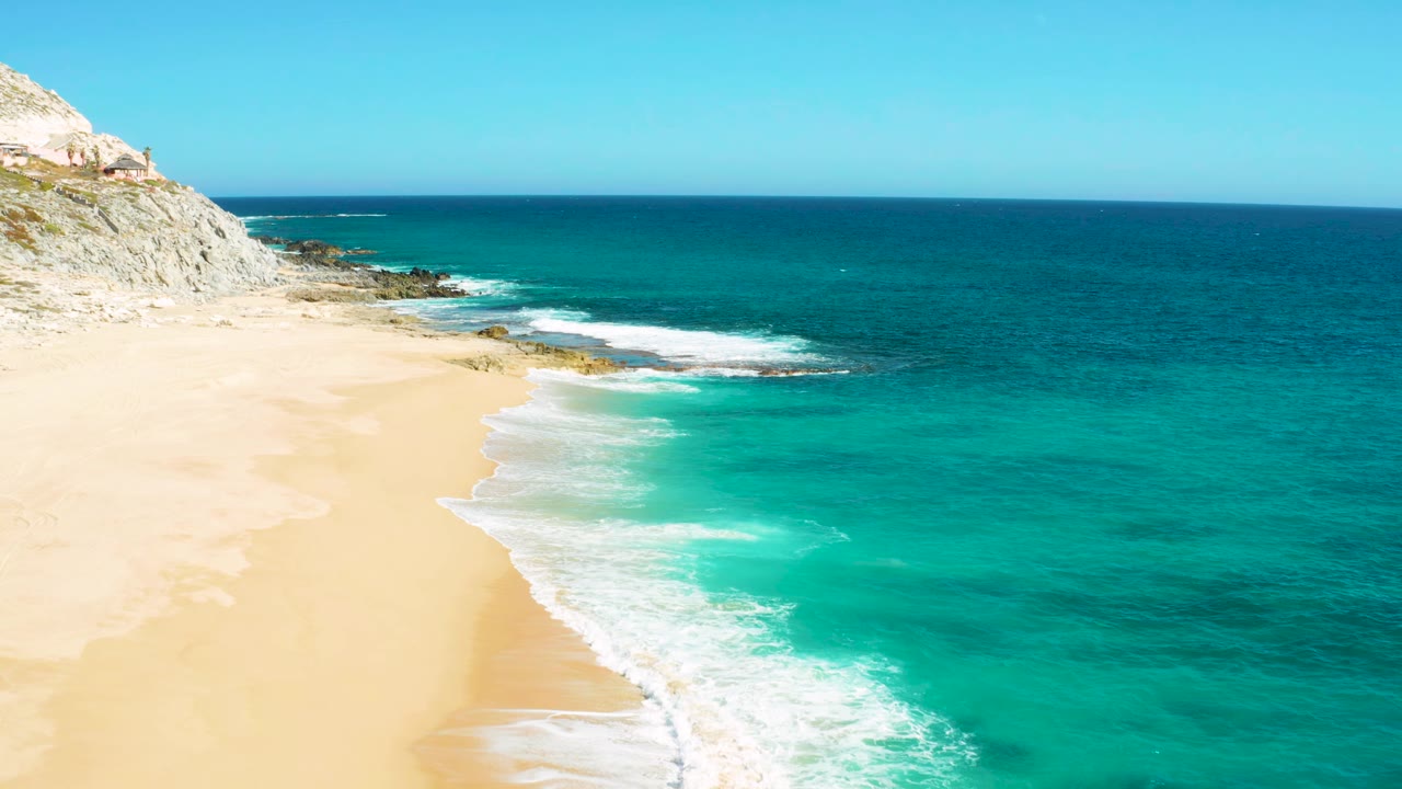 Aerial frame from East Cape Baja California Sur, Mexico, with waves curling farther up the sandy shoreline, rocky outcrops near the upper left coast, open turquoise water, and no visible people.