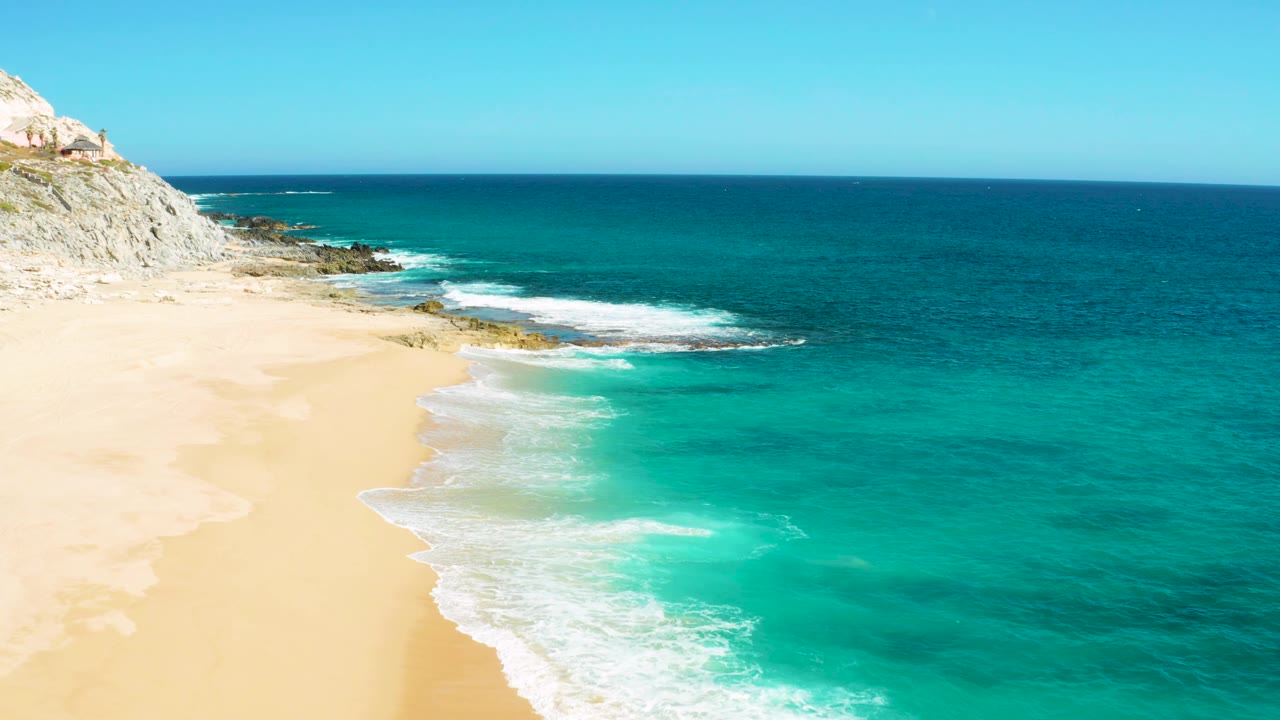 Aerial frame from East Cape Baja California Sur, Mexico, showing more open turquoise sea to the right, a sandy beach on the left, surf breaking near a rocky point, and no visible people.