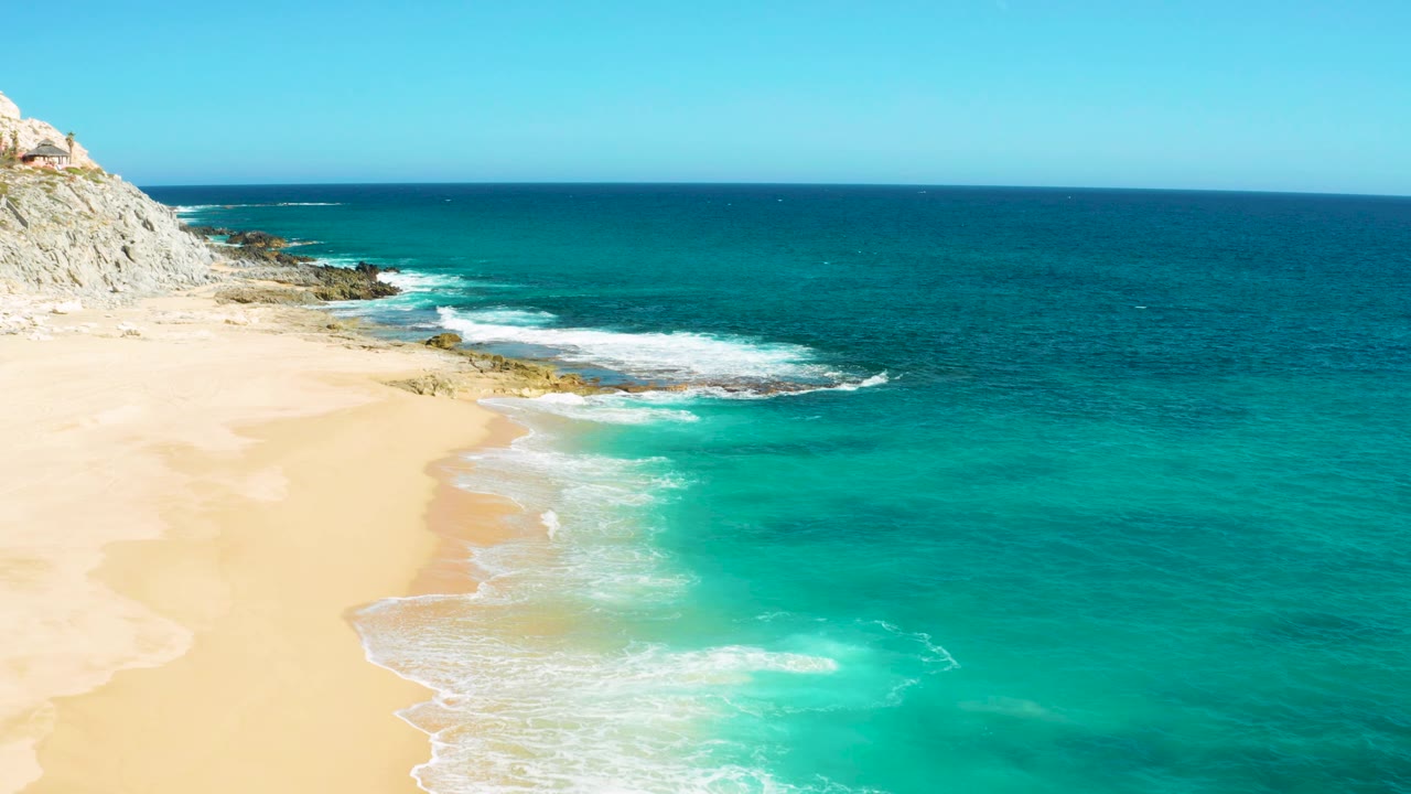 Aerial frame from East Cape Baja California Sur, Mexico, where a foamy wave spreads along the pale sand, the rocky coast sits at upper left, turquoise water fills the right side, and no visible people appear.