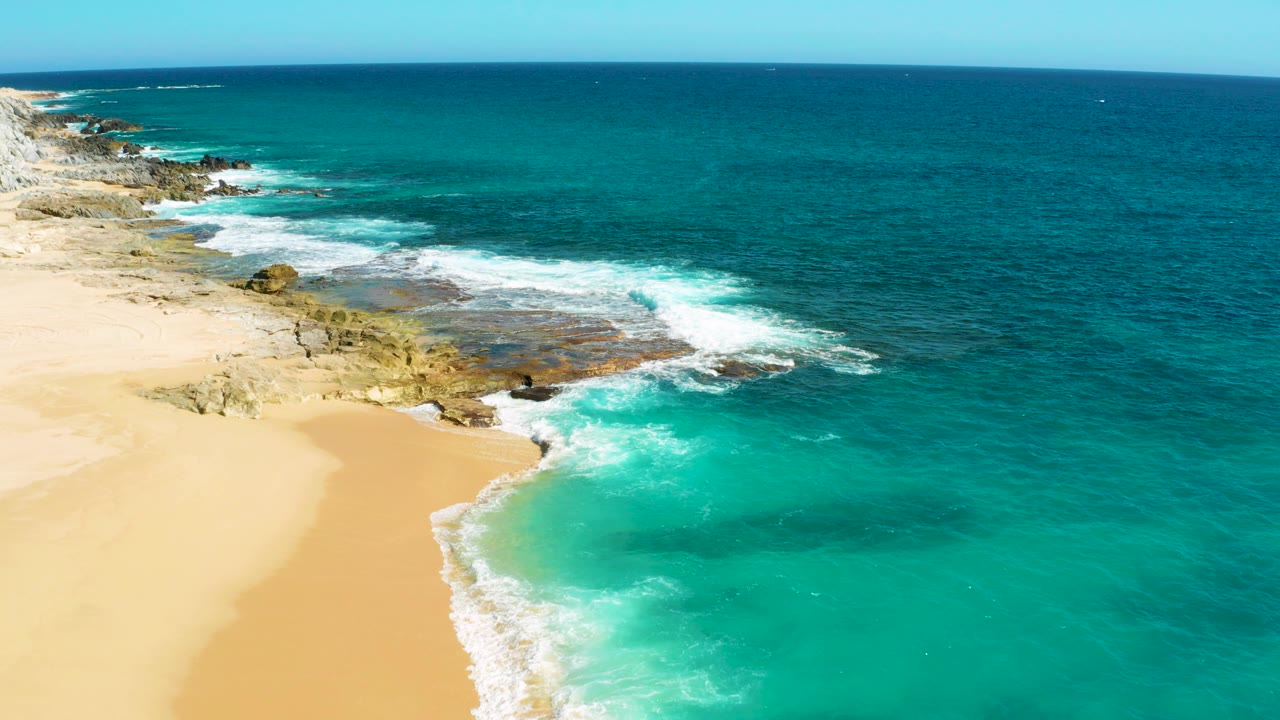 Aerial frame from East Cape, Baja California Sur showing a sandy beach at lower left, tan rock ledges along the shore, turquoise surf, deep blue ocean, and no visible people.