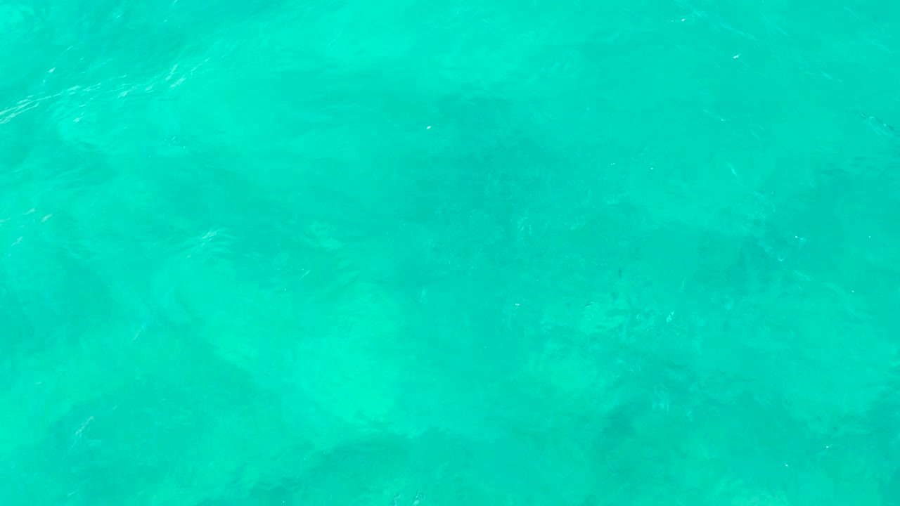 Blue-green ocean surface in East Cape, Baja California Sur, with small ripple lines crossing the frame and muted seabed texture visible below, no visible people.
