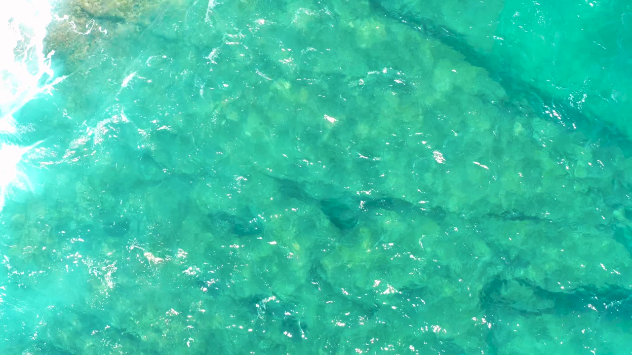 Overhead East Cape water frame with blue-green shallows, scattered underwater rock texture, and a pale wash of wave foam entering from the left edge, no visible people.