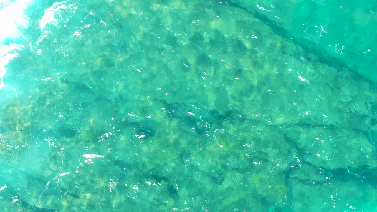 Clear turquoise ocean in East Cape, Baja California Sur, seen from above with darker rock channels under the surface and small white highlights on the ripples, no visible people.