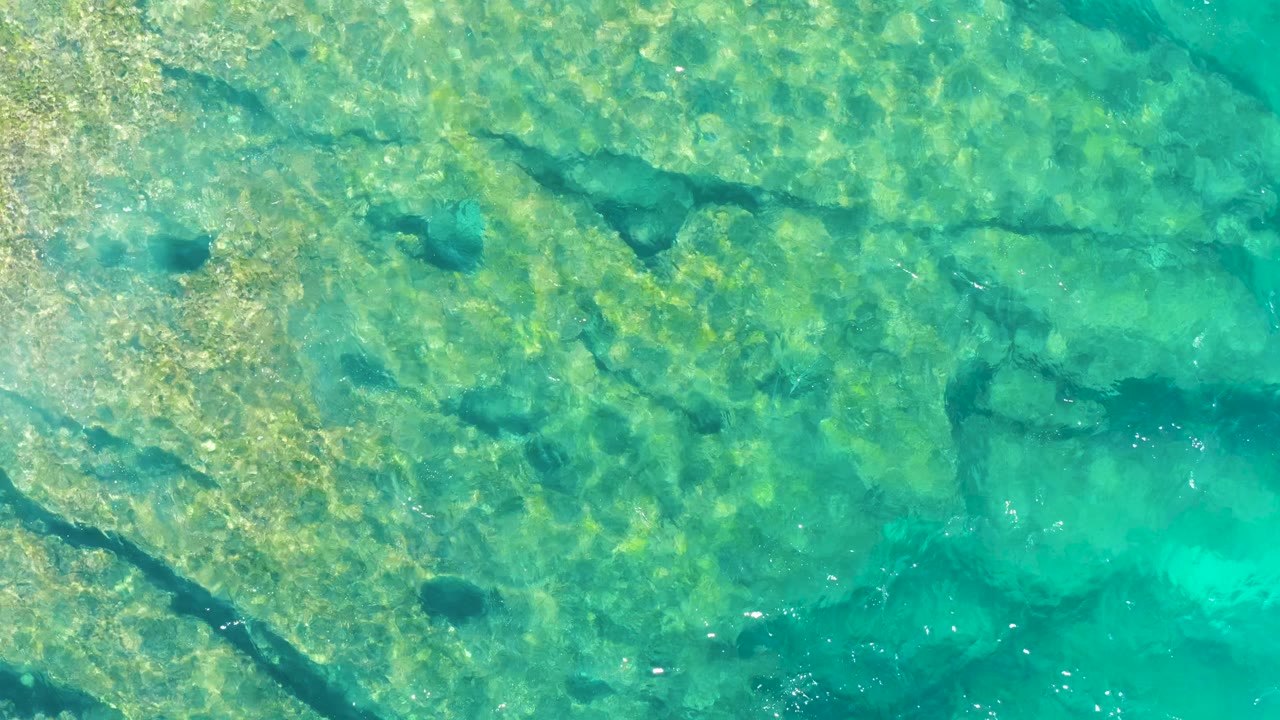 Turquoise water covers a rocky seabed in East Cape BCS Mexico, with dark natural channels cutting across the upper half and no visible people.