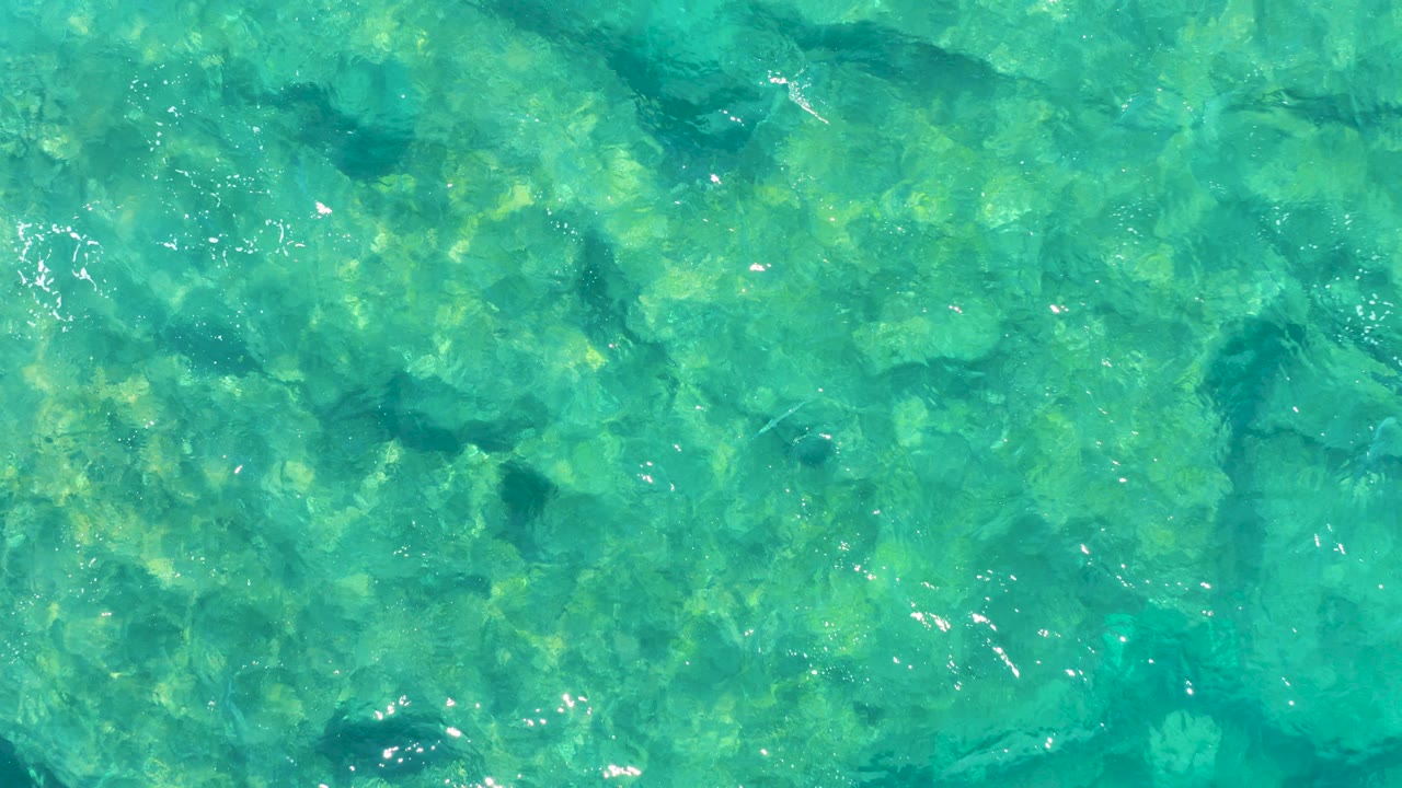 Straight-down aerial view of clear turquoise water over shallow coral and rocks in East Cape BCS Mexico, with no visible people.