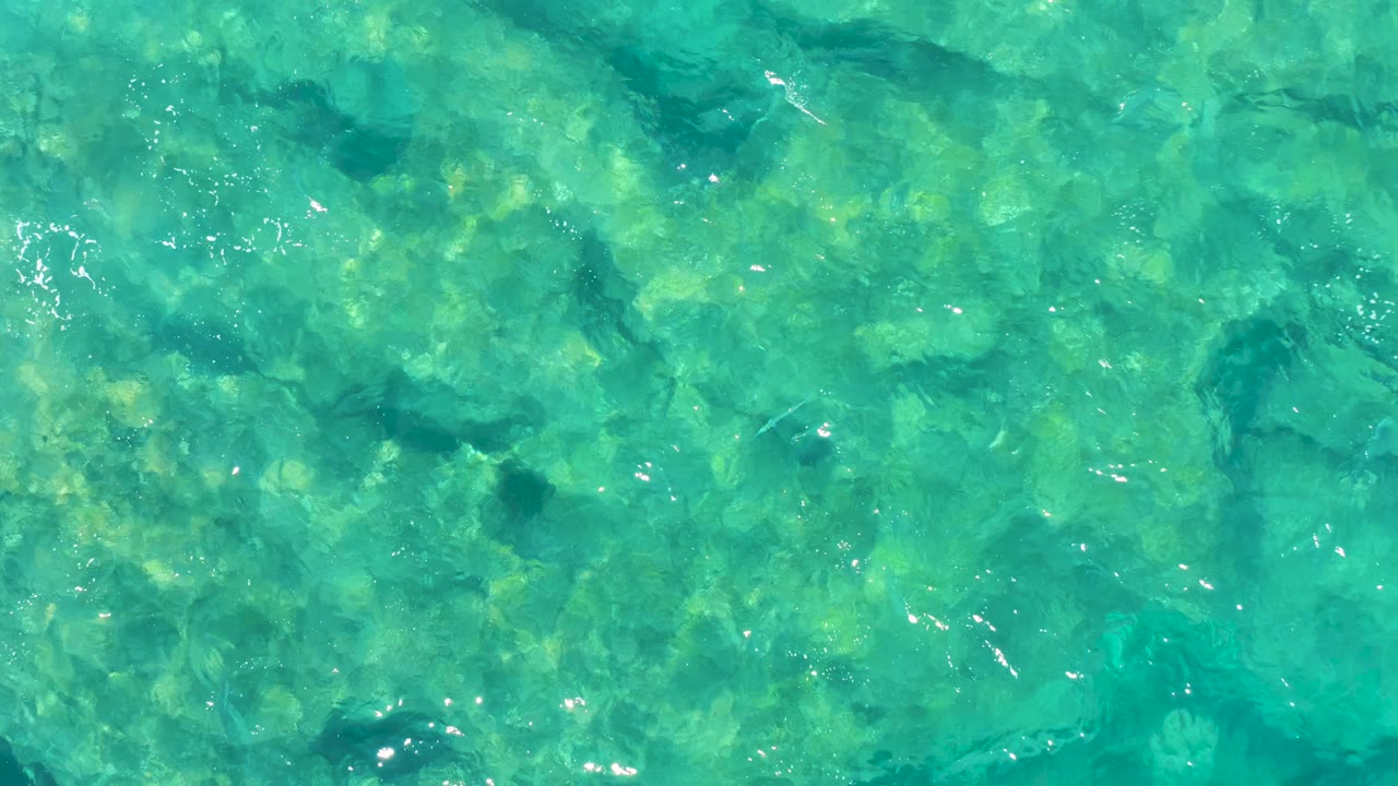 Green-blue rippled ocean surface reveals mottled coral and rock shapes below in East Cape BCS Mexico, with no visible people.
