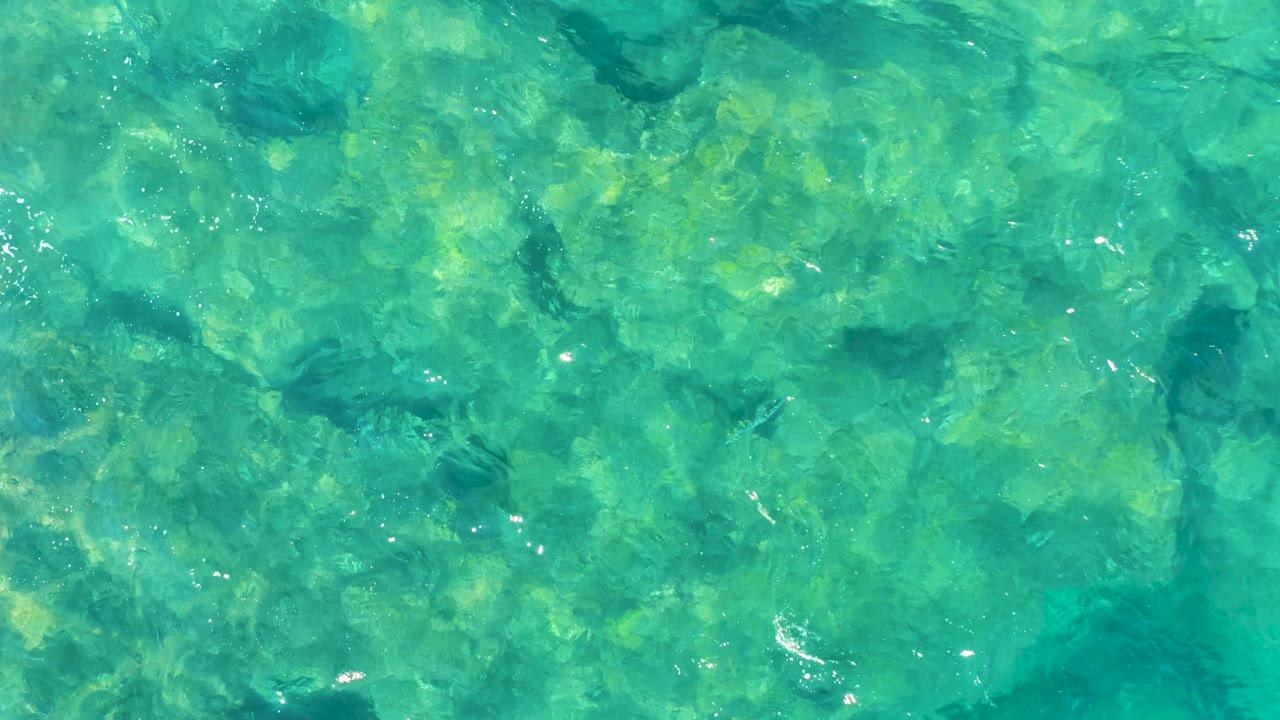Clear shallow ocean water shows darker coral patches and pale rock areas under a turquoise surface in East Cape BCS Mexico, with no visible people.