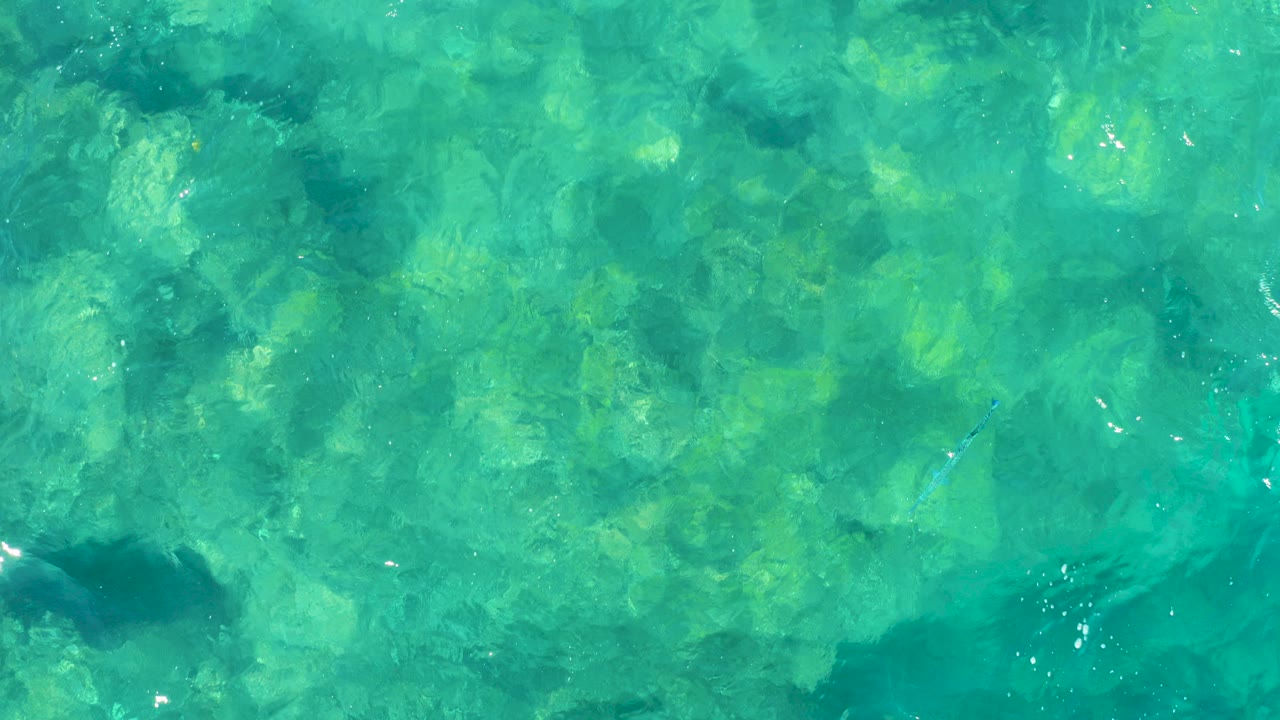 Overhead view of clear turquoise water in East Cape, Baja California Sur, Mexico, with submerged rock shapes, faint ripples, bright reflections, and no visible people.