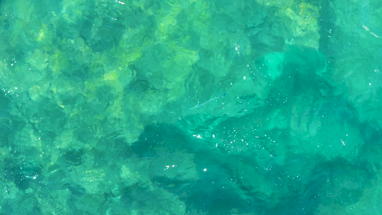 Aqua shallow water in East Cape, Baja California Sur, seen from above with darker submerged patches, pale rock areas, fine ripples, and no visible people.