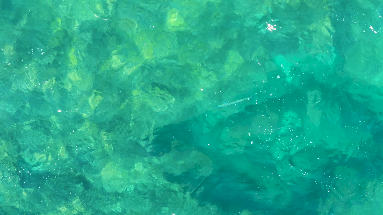Overhead view in East Cape, Baja California Sur, where turquoise water reveals a rocky bottom, shifting surface texture, and no visible people.