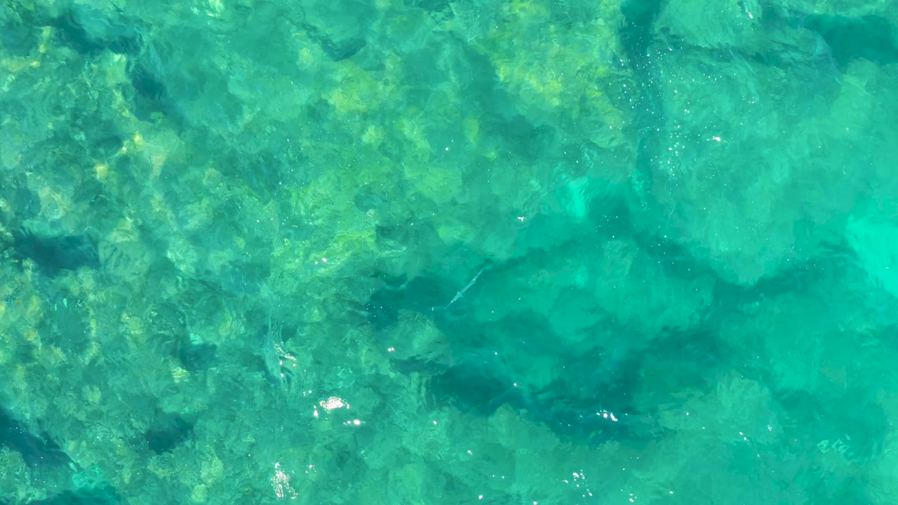 Clear blue green ocean water in Baja California Sur shows darker submerged rock shapes, mild ripples, scattered light reflections, and no visible people.