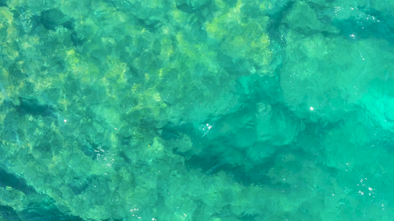 A top-down Baja California Sur water view shows turquoise shallows, mottled rocky seabed texture, bright surface sparkles, and no visible people.