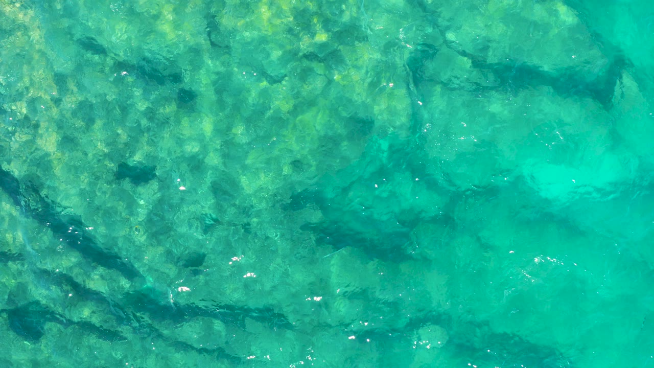 Overhead view of clear turquoise water over a rocky seabed in East Cape, Baja California Sur, with small sun glints near the surface and no visible people.