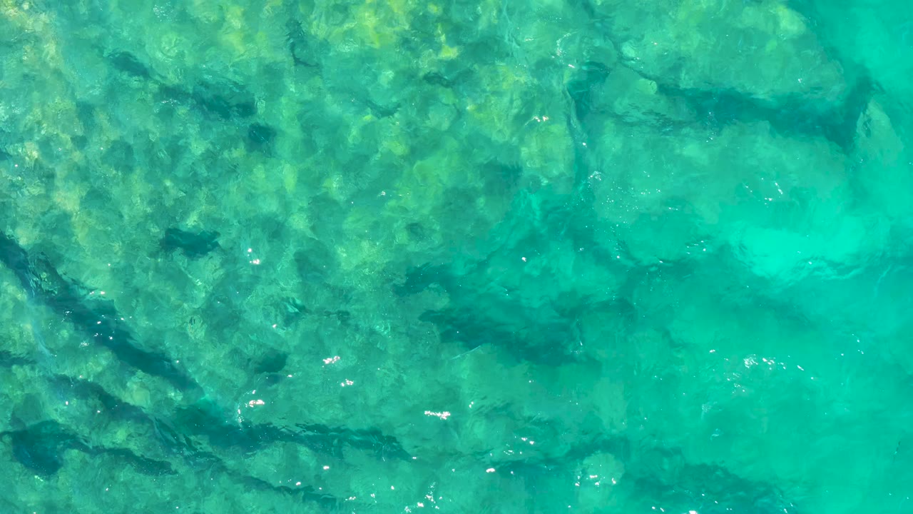 Clear turquoise ocean water in East Cape, Baja California Sur, shows darker rock lines beneath the surface, scattered light flecks, and no visible people.