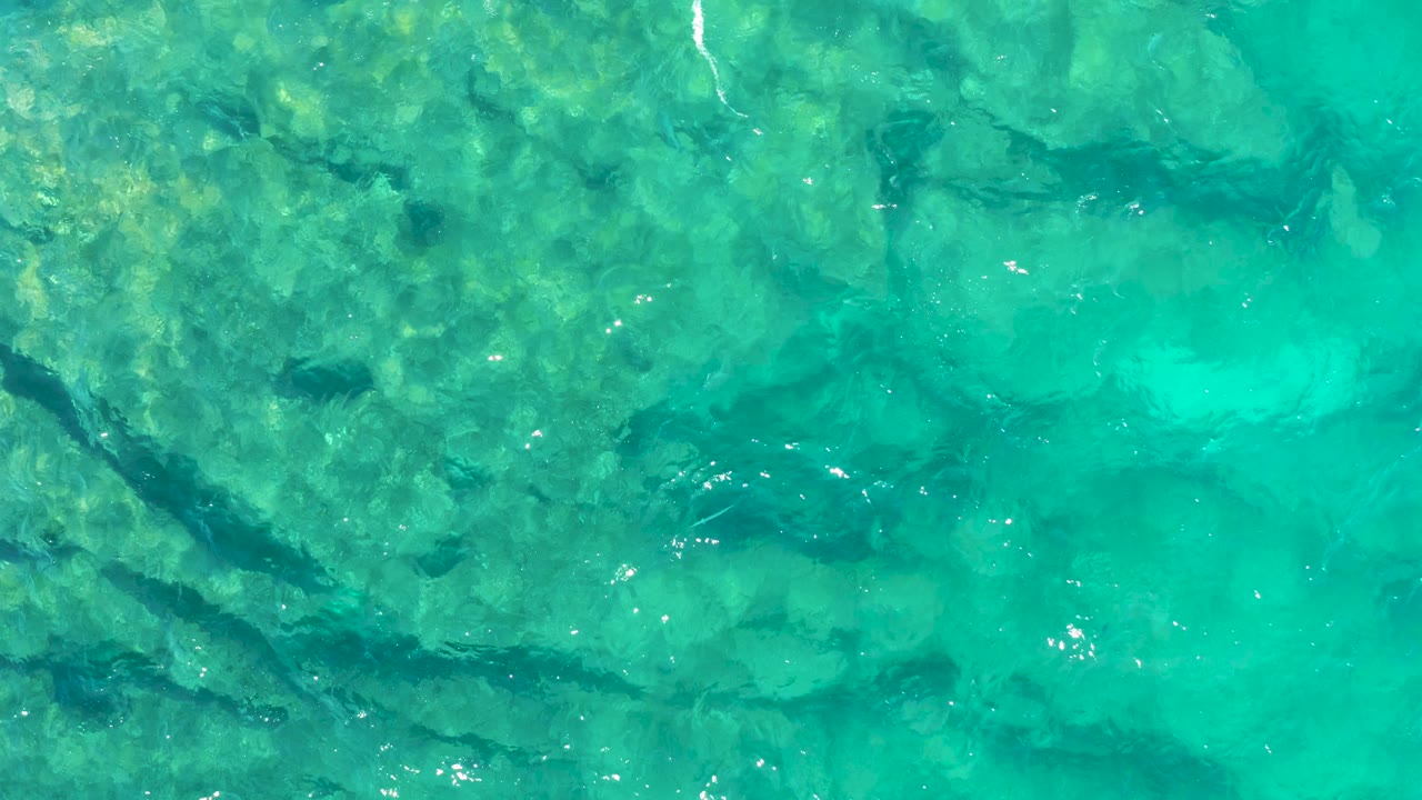 Aerial-looking frame of shallow turquoise water in East Cape, Baja California Sur, with pale rock patches, darker underwater cracks, surface sparkle, and no visible people.