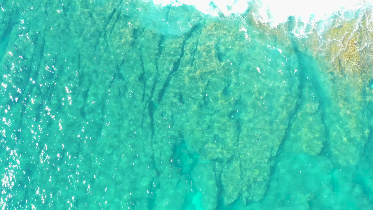 Straight-down drone frame over East Cape BCS Mexico shallows, with white foam pushing across the upper right and rock shapes visible under the turquoise water with no visible people.