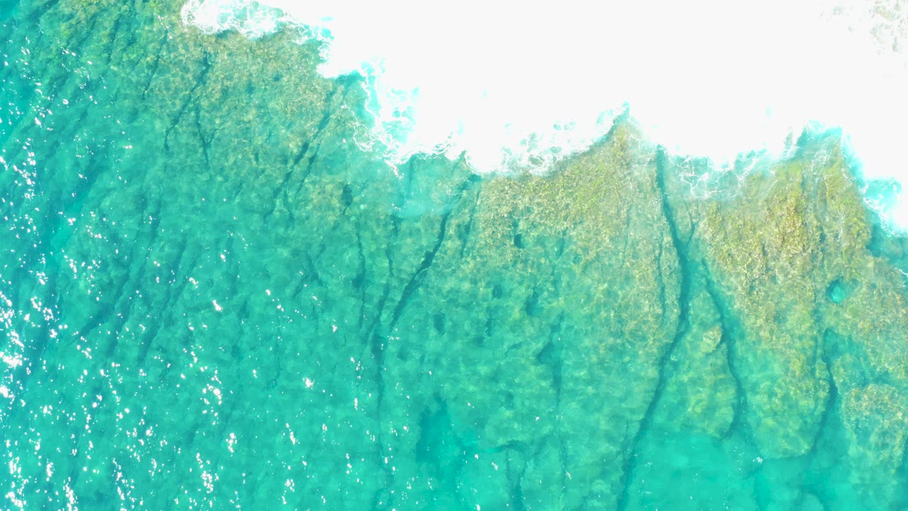 Top-down view of turquoise water over a textured seabed in East Cape BCS Mexico, with white wave foam along the upper edge and no visible people.