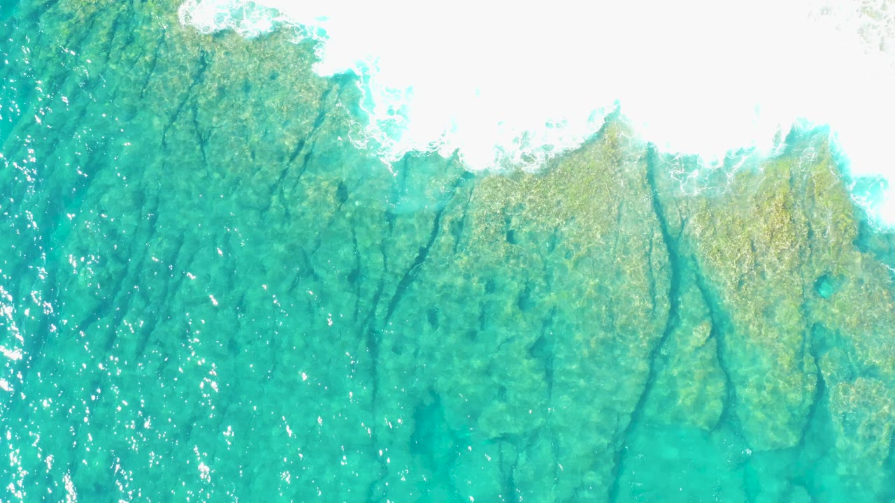 Overhead frame of clear blue-green ocean in East Cape BCS Mexico, with branching dark seabed marks and bright foam near the top, no visible people.