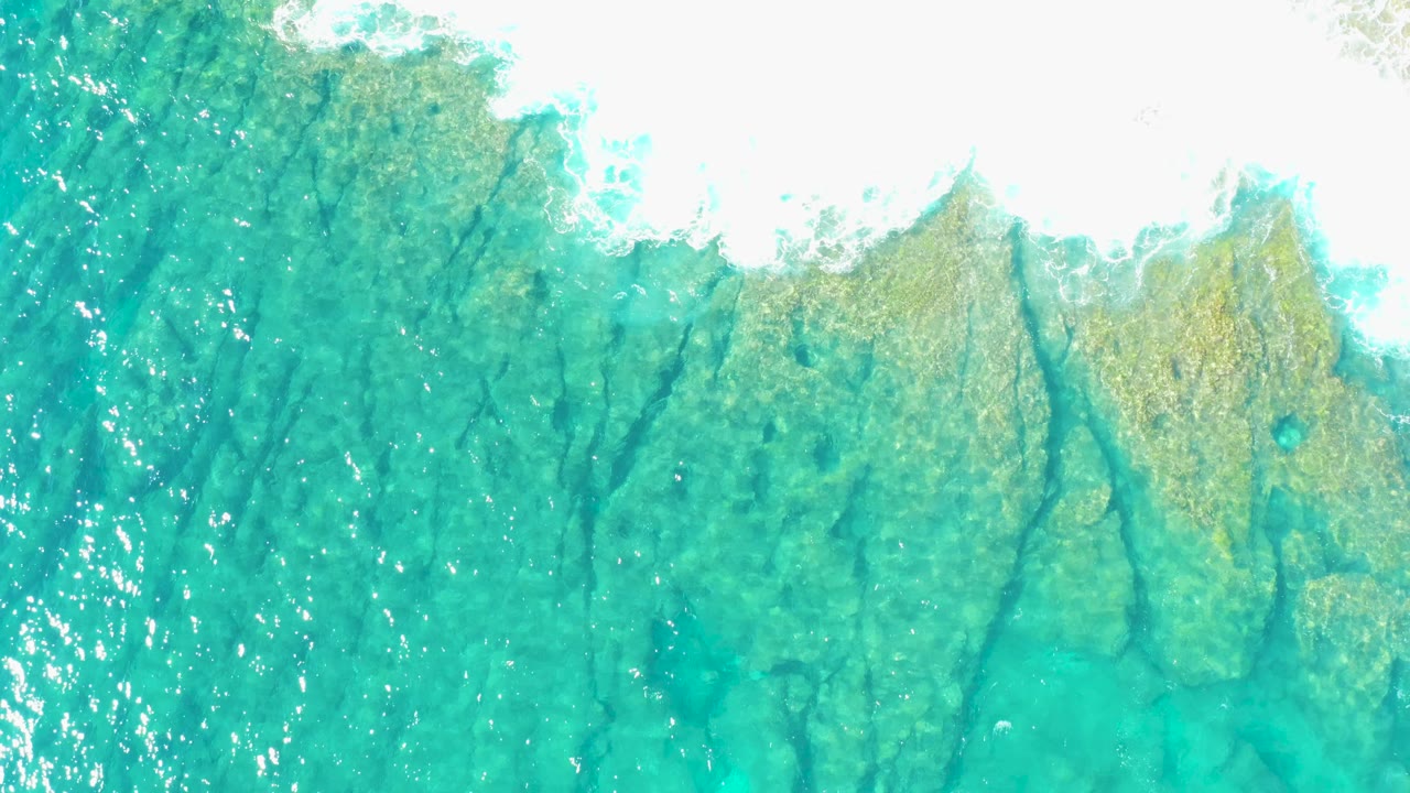 Top-down aerial of turquoise shallows in East Cape BCS Mexico, where mottled seabed color shows through the water and white surf foam borders the upper frame, no visible people.