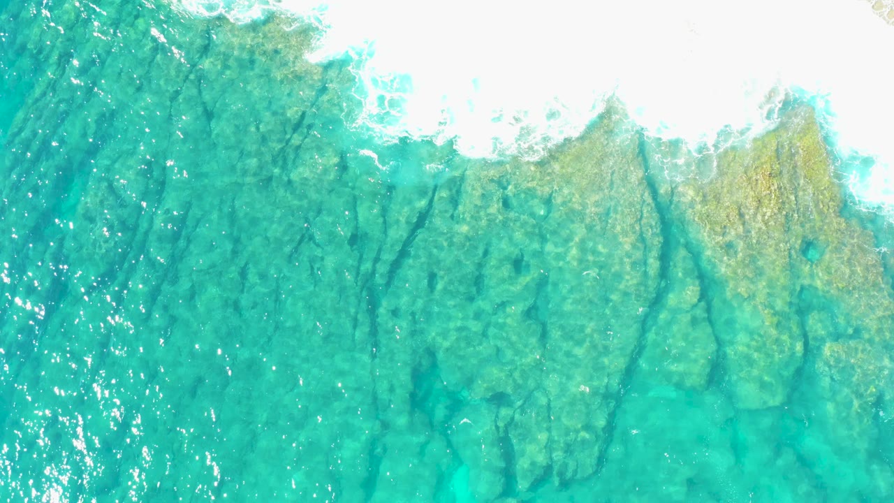 Foam washes across the top of clear turquoise water above rocky seabed texture.