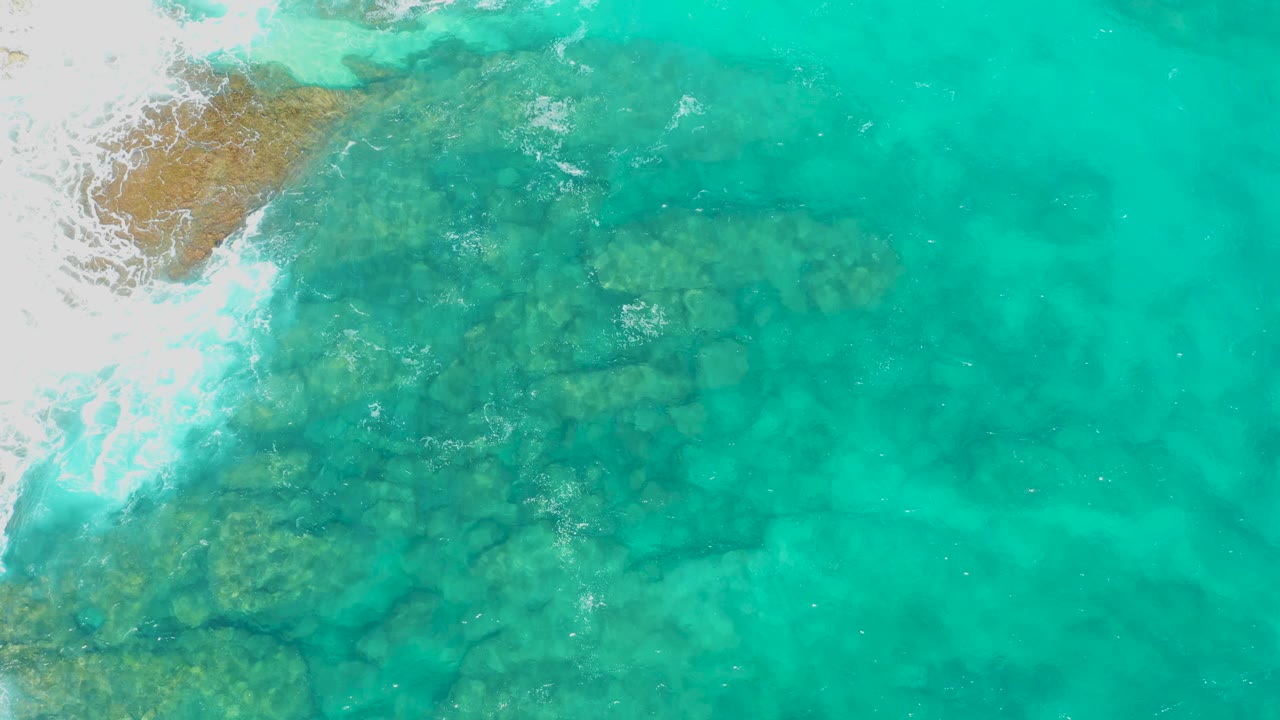Aerial frame in Baja California Sur showing clear turquoise water over a broad rocky patch, with foamy wave wash along the left side and no visible people.