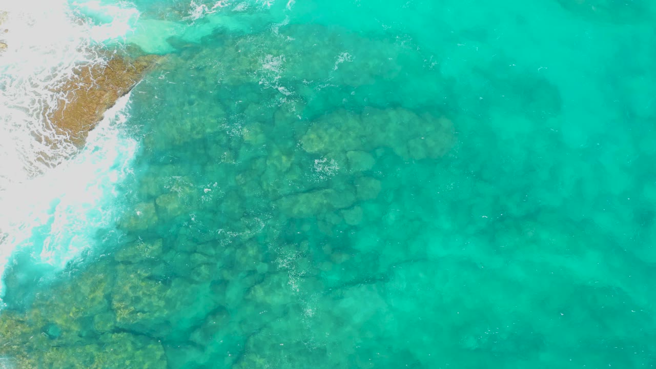 Aerial view in Baja California Sur with a rocky seabed centered under turquoise water, white breaking surf near the upper left corner, and no visible people.