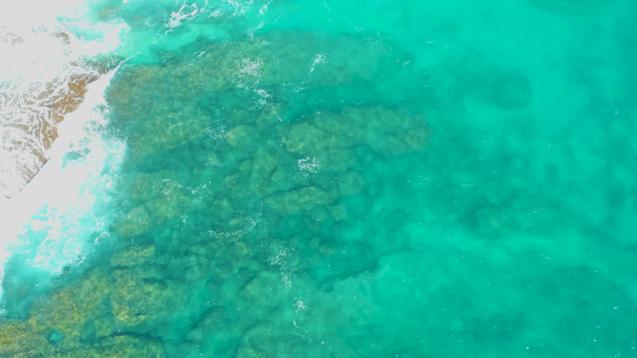 Aerial frame in Baja California Sur where bright white surf pushes in from the left beside darker submerged rocks and clear turquoise water, with no visible people.