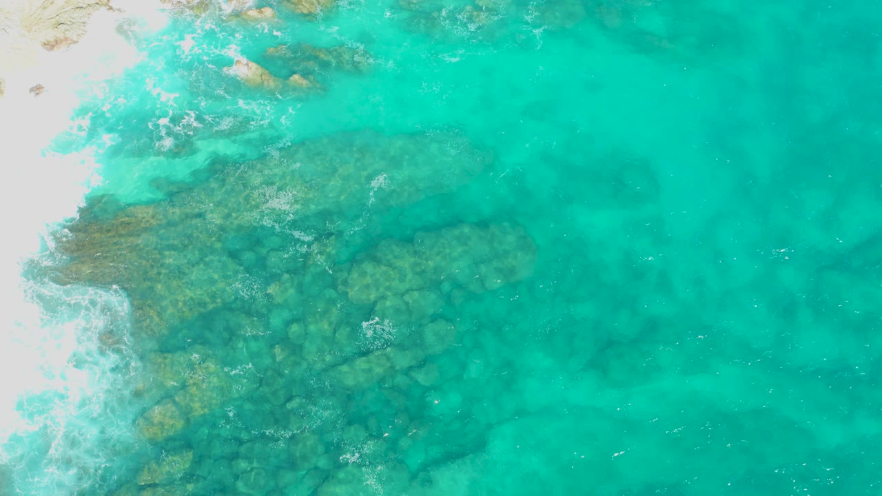 Overhead turquoise shallows with submerged rocks and white surf at the sand edge.