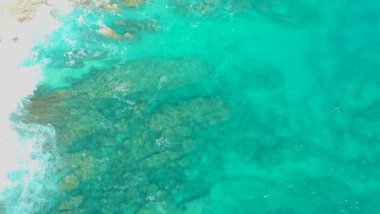 Aerial frame of clear turquoise water revealing rocks below the surface, with white sand and surf foam along the shoreline in East Cape, Baja California Sur, Mexico, and no visible people.