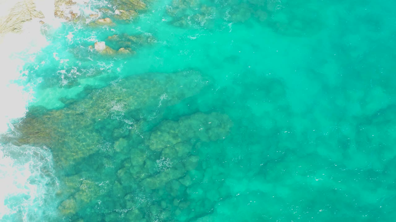 Top-down frame of bright turquoise shallows over uneven rock patches, with white sand and foam near the upper left in East Cape, Baja California Sur, Mexico, and no visible people.