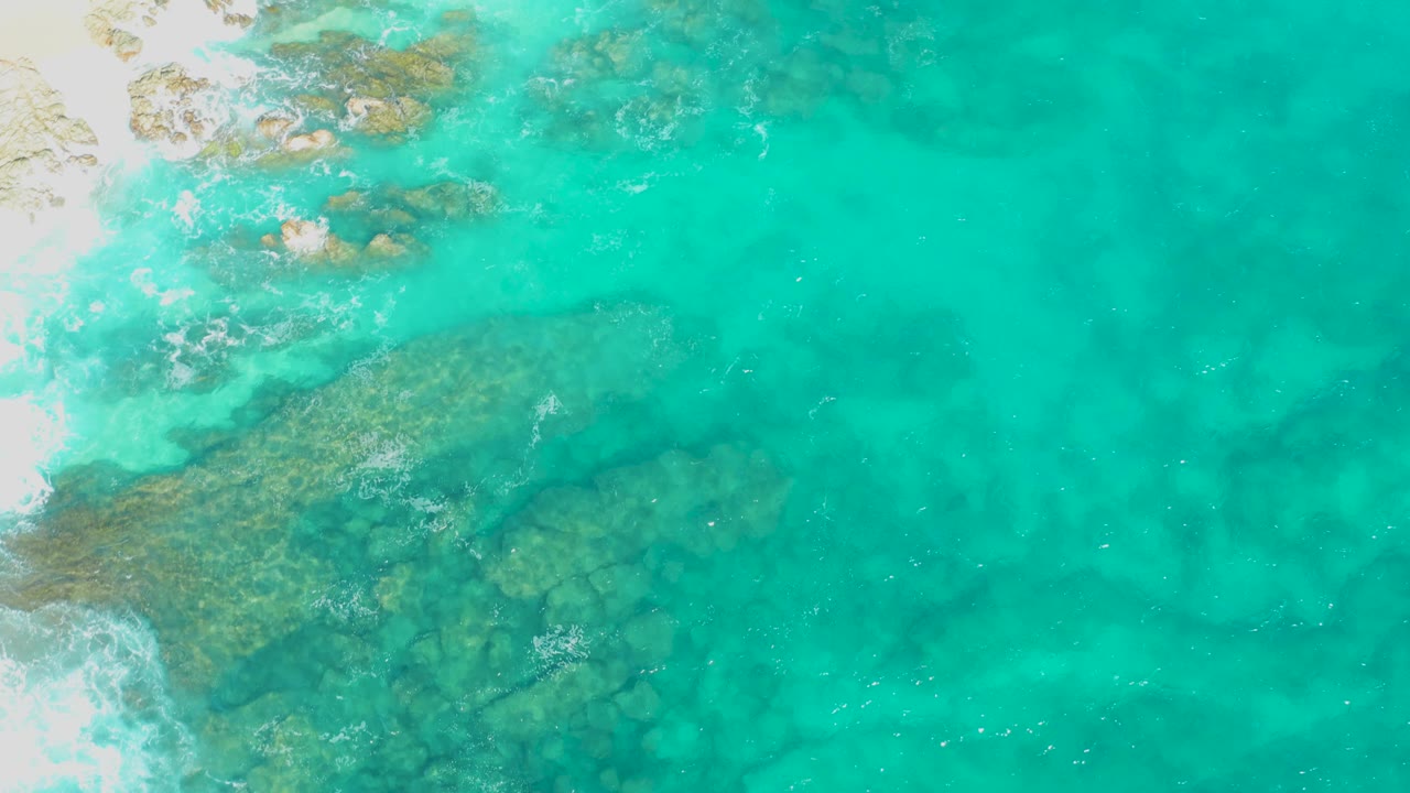 Aerial view of turquoise coastal water with a broad submerged rocky area and a foamy white sand edge entering from the left in East Cape, Baja California Sur, Mexico, with no visible people.