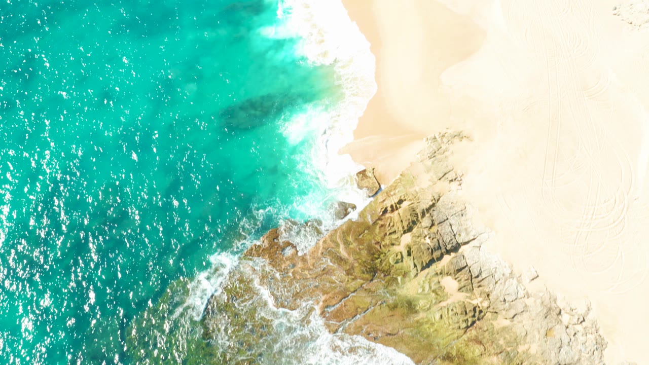 Overhead view of turquoise water, white foam, pale sand, and rocky outcrops on the East Cape coastline in Baja California Sur, Mexico, with no visible people.