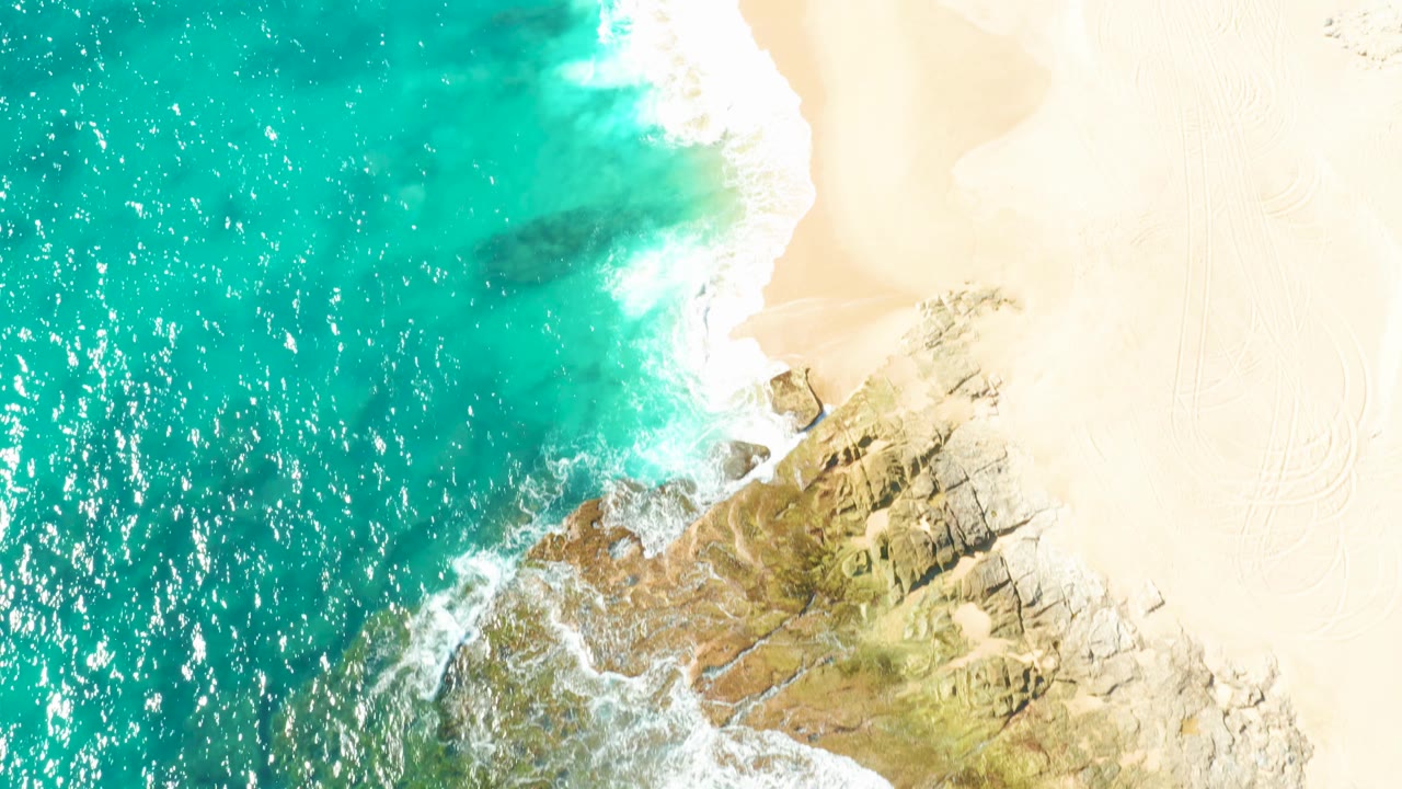 Aerial frame of clear turquoise ocean pushing white foam against tan rocks and pale beach on the East Cape shore, with no visible people.