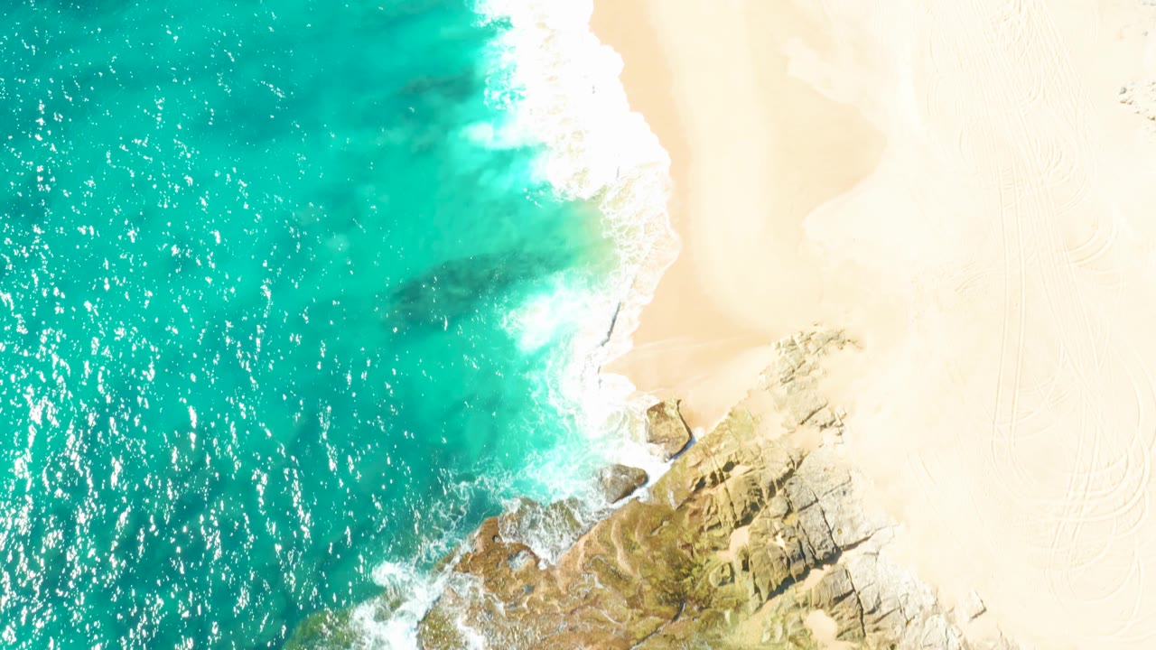 Top-down East Cape coastal view with sparkling turquoise water on the left, white surf at the beach edge, and rocks extending into pale sand, with no visible people.