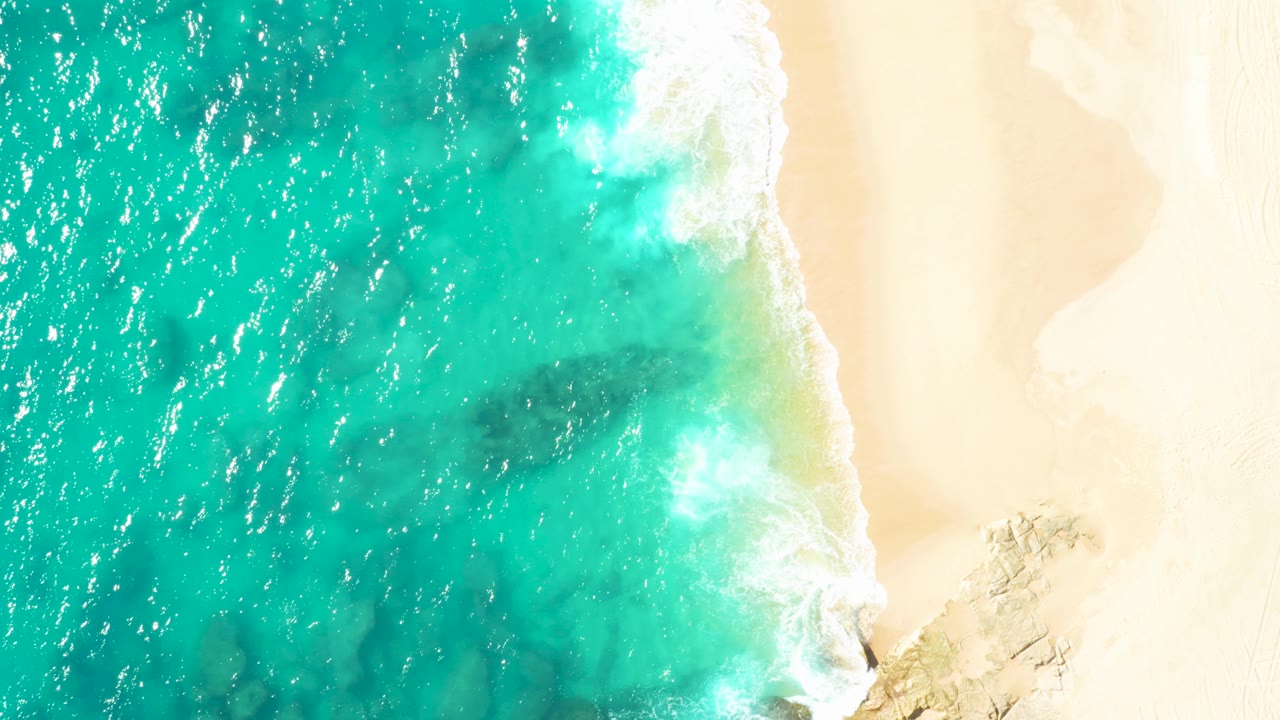 Top-down aerial view of turquoise water meeting pale sand in Baja California Sur, Mexico, with white foam near a rocky edge and no visible people.